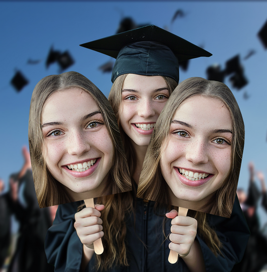 A newly graduated student proudly holds a giant cutout of her own face while celebrating at her graduation ceremony, smiling with excitement.