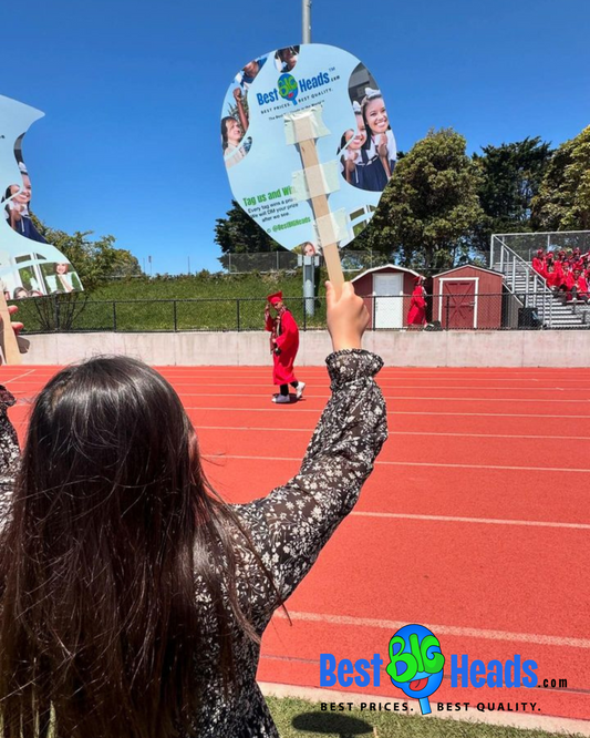 A photo of a proud mom enthusiastically waving a big head cutout of her son’s face as he marches during the graduation ceremony, beaming with pride and joy.