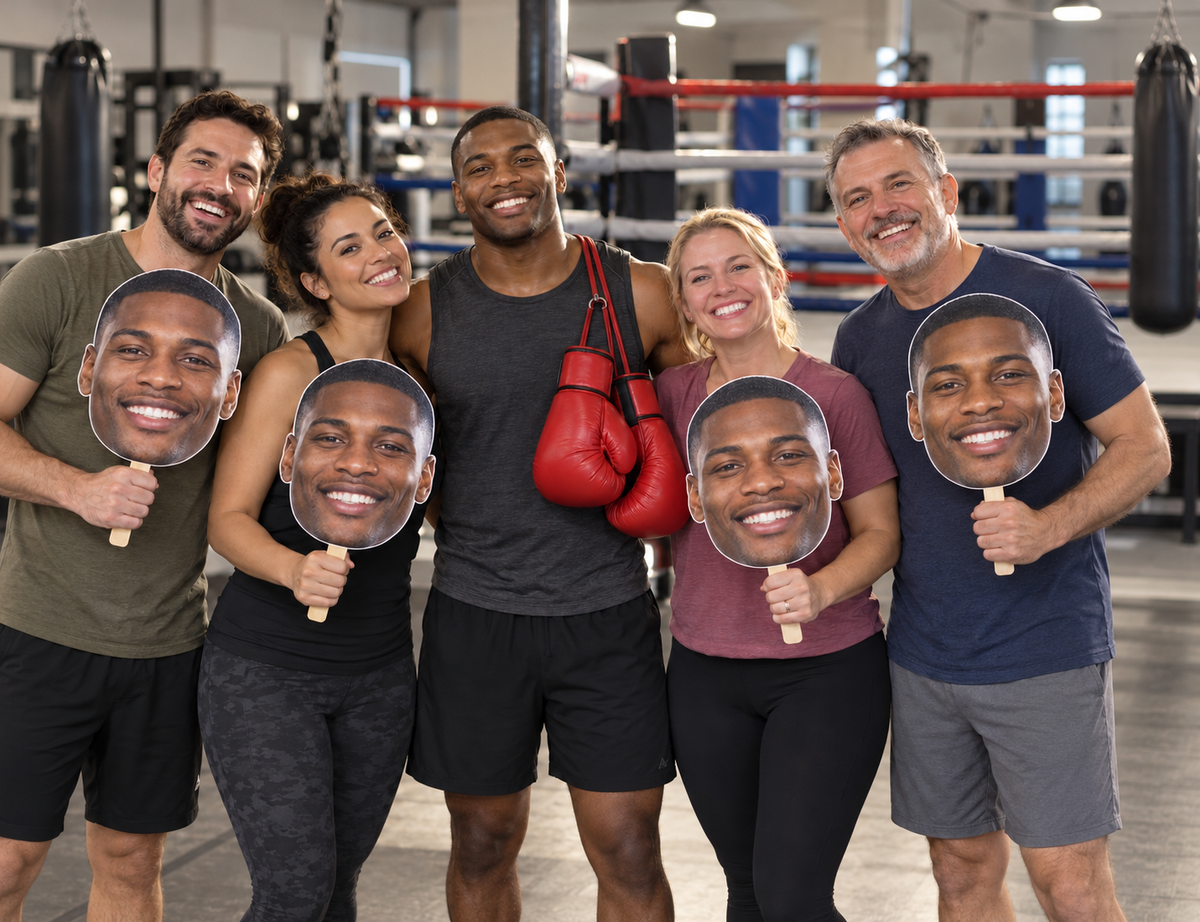 Five adults in a boxing gym, with four supporters holding oversized face-only cutouts of the smiling boxer wearing red gloves around his neck.