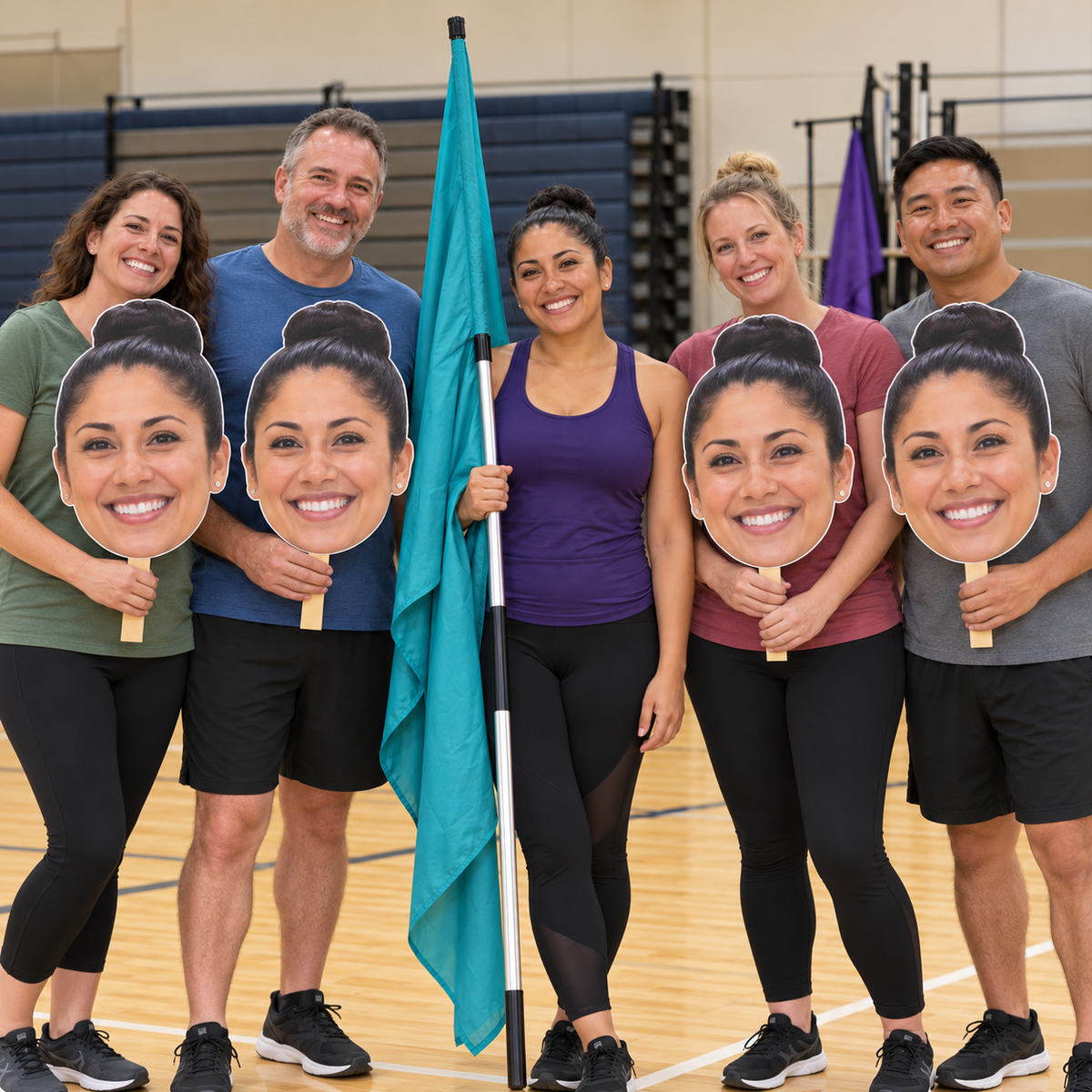 Five smiling adults in a gym, with four holding matching oversized face cutouts on flat-ended wooden handles beside a woman holding a turquoise color guard flag at rest.